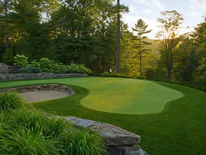 A professionally installed backyard putting green featuring smooth green turf, white cup, and surrounding landscape pavers.